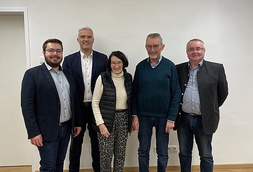 Vier Männer und eine Frau stehen in einer Gruppe in einem Raum und schauen in die Kamera.