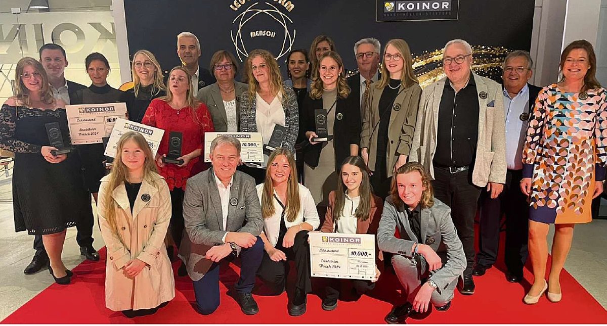 Eine große Gruppe mit Männern und Frauen in verschiedenen Altersklassen stehen auf einem roten Teppich. Die Personen in der ersten Reihe knien. Sie alle lächeln in die Kamera. 
