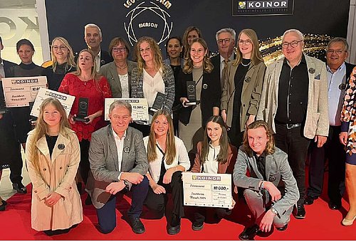 Eine große Gruppe mit Männern und Frauen in verschiedenen Altersklassen stehen auf einem roten Teppich. Die Personen in der ersten Reihe knien. Sie alle lächeln in die Kamera. 