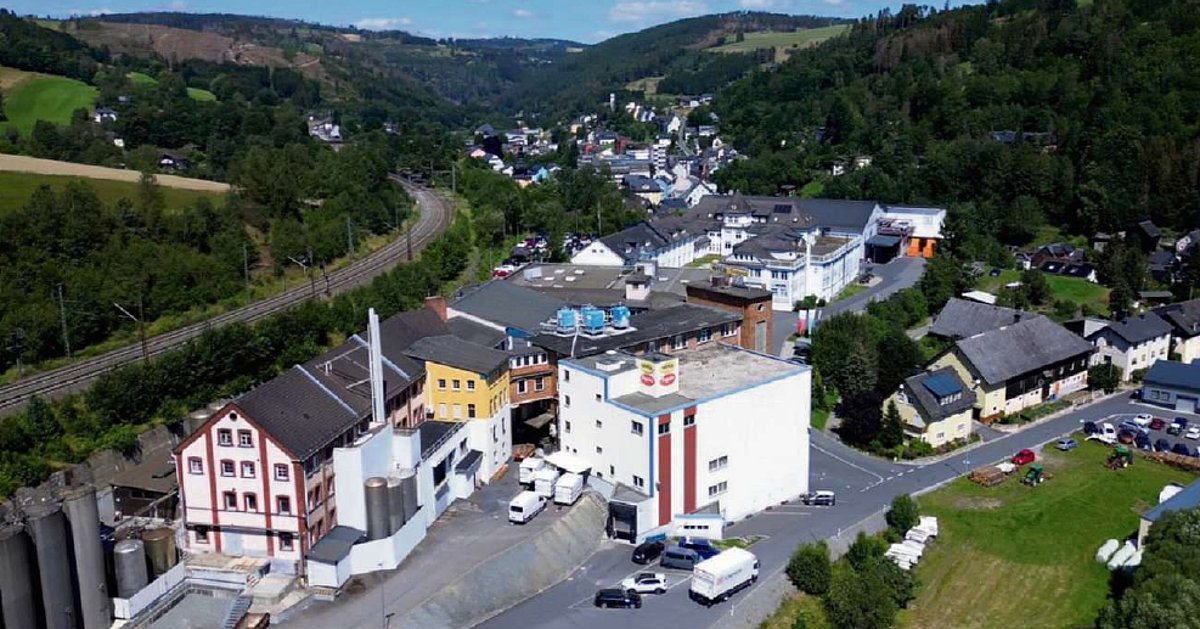 Luftbild einer Stadt. Im Hintergrund sind Berge, teilweise mit Waldbewuchs. Im Vordergrund sind mehrere Industriegebäude zusehen. Es stehen Autos auf den Parkplätzen. Dahinter sieht man Wohnhäuser.