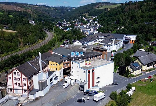 Luftbild einer Stadt. Im Hintergrund sind Berge, teilweise mit Waldbewuchs. Im Vordergrund sind mehrere Industriegebäude zusehen. Es stehen Autos auf den Parkplätzen. Dahinter sieht man Wohnhäuser.
