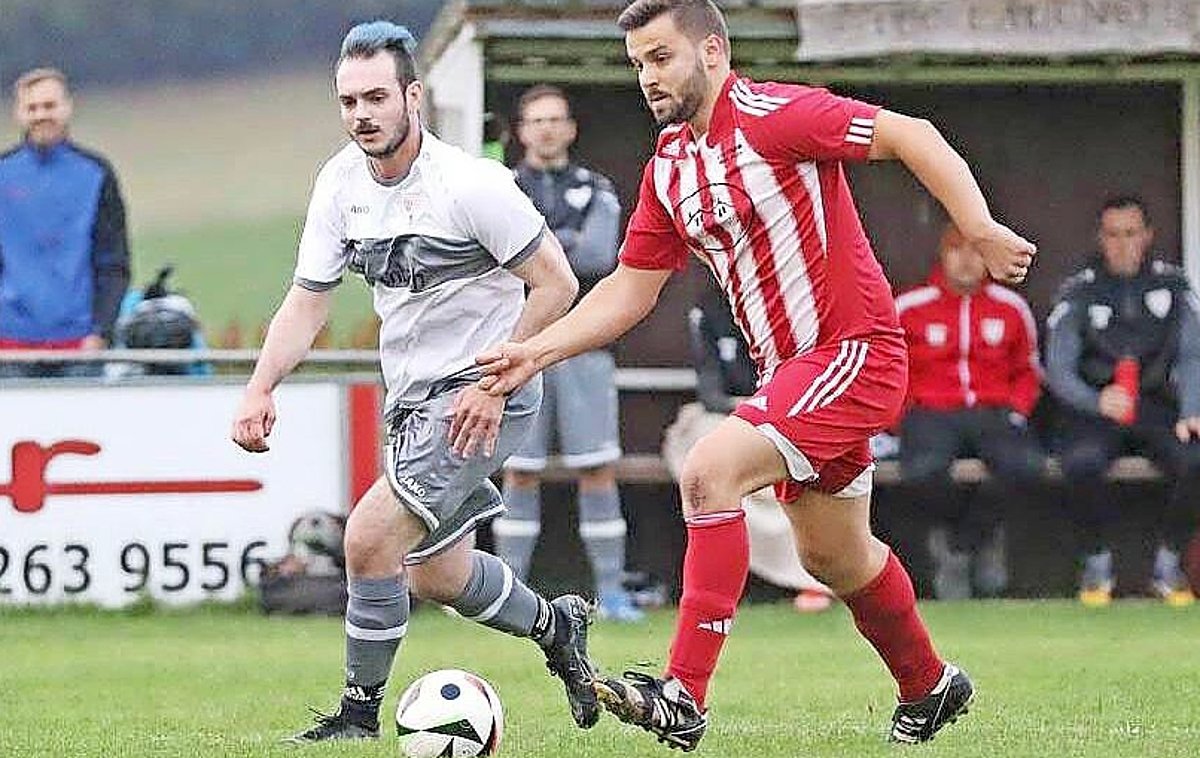Zwei Männer auf einem grünen Rasen mit einem Fußball. Der eine trägt ein rotes Trikot, der andere Spieler ein weißes Trikot.