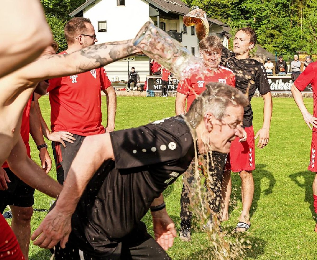 Auf einem Fußballplatz stehen mehrere Männer in roten Trikots in einer Runde. In ihrer Mitte ein Mann in schwarzem Trikot. Über seinem Kopf wird gerade ein Glaskrug mit Bier ausgeschüttet.