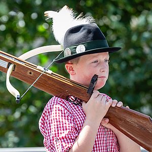 Eine Junge mit rot-weiß-kariertem Hemd und einem Filzhut mit langen weißen Federn trägt eine hölzerne Armbrust auf seiner Schulter. Er schaut nach links.