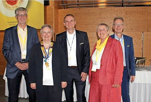 Drei Männer und zwei Frauen in Businesskleidung stehen in einer Gruppe zusammen. Sie stehen in einem Gebäude und lächeln in die Kamera.