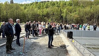 Auf einem Baustellengelände stehen viele Menschen. Sie schauen auf einen Mann, der etwas erklärt.