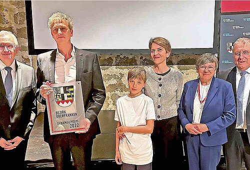 Im Foto zu sehen sind der Geehrte mit seinem Sohn und seiner Frau. Umrahmt werden Sie von dem Bezirksheimatpfleger, der Regierungspräsidentin und dem Bezirkstagspräsidenten. Alle Personen sind festlich gekleidet.