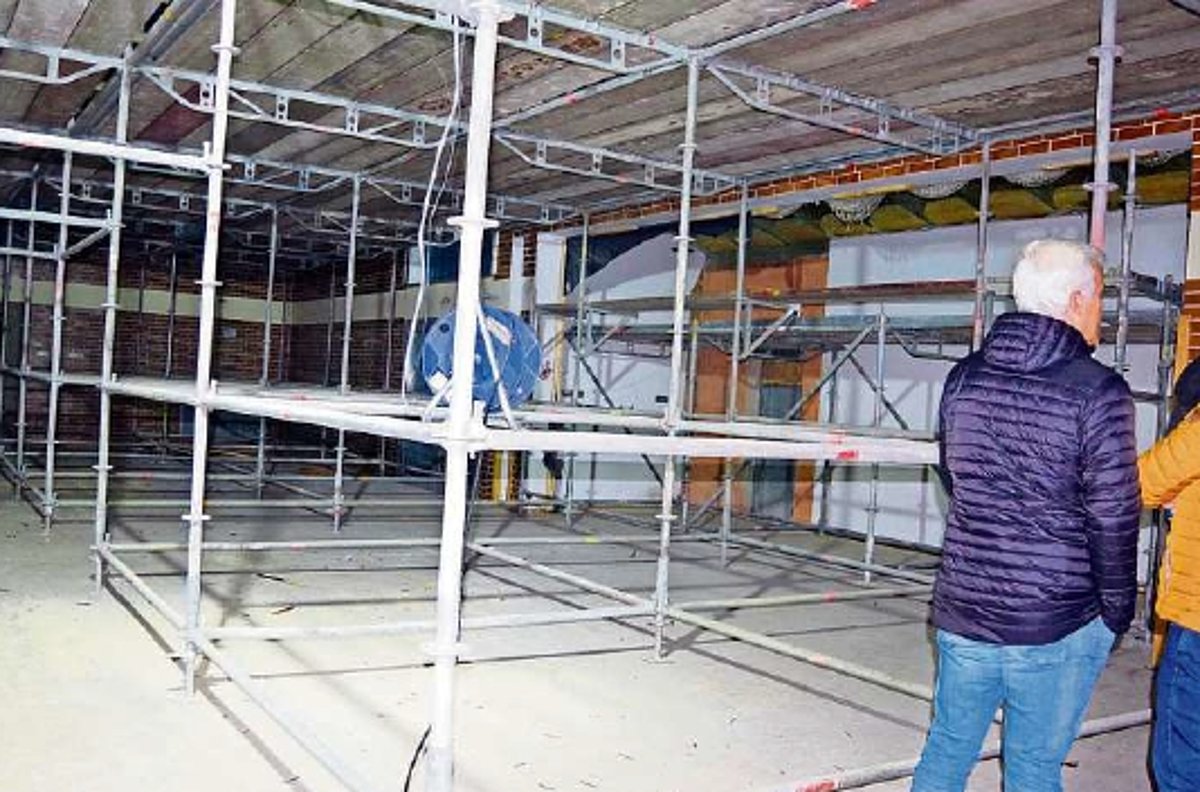 Ein großes Baugerüst in einer Halle. Am Rand sind zwei Personen zu sehen, die sich die Baustelle ansehen.