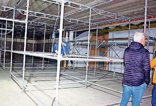 Ein großes Baugerüst in einer Halle. Am Rand sind zwei Personen zu sehen, die sich die Baustelle ansehen.