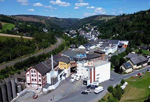 Neue und alte Betriebsgebäude mit bewaldeten Bergen rundherum aus der Luft fotografiert.
