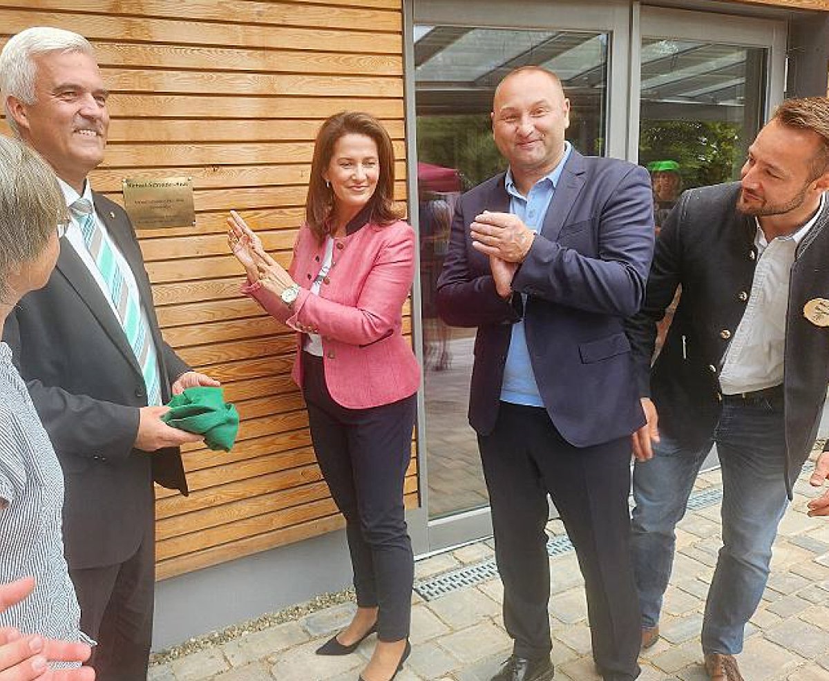 Drei Männer in Anzügen und zwei Frauen, eine davon Staatsministerin Michaela Kaniber, stehen vor einem Gebäude mit Holzverkleidung. Frau Kaniber deutet auf ein Schild an der Hauswand. Alle lächeln.