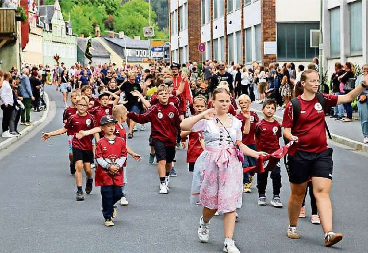 Ein buntes Bild mit vielen Zuschauern links und rechts am Straßenrand. Im Vordergrund eine Gruppe von Mädchen und Jugen im Fußballdress, angeführt von einer Trainerin im Begleitung eines Mädchens im Dirndl.
