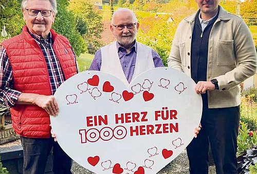 Drei Männer stehen nebeneinander und halten ein großes Herz aus Pappe. Alle drei schauen in die Kamera
