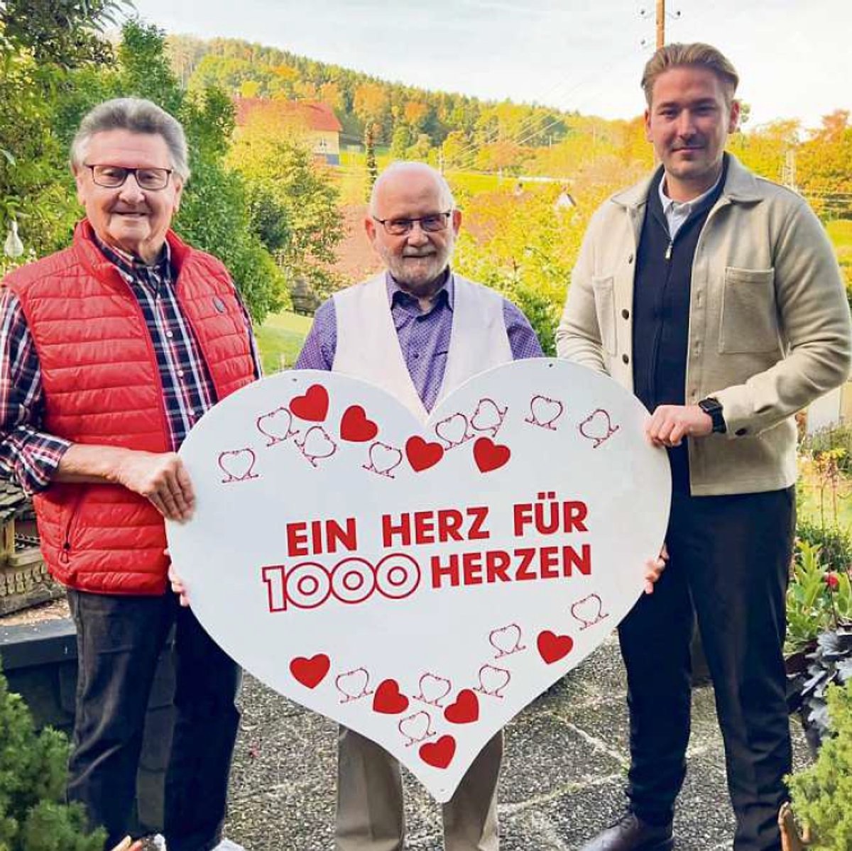 Drei Männer stehen nebeneinander und halten ein großes Herz aus Pappe. Alle drei schauen in die Kamera