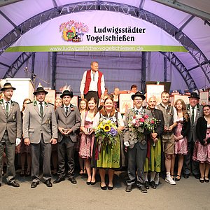 In einem Festzelt vor einer große Bühne steht eine Gruppe von Menschen. Die Männer tragen Schützenuniformen, die Frauen tragen Dirndl. Die Personen in der Mitte halten je einen Pokal in ihren Händen.