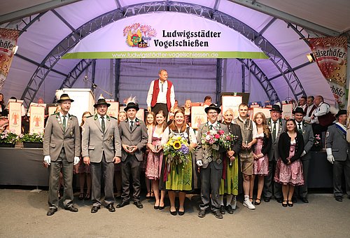 In einem Festzelt vor einer große Bühne steht eine Gruppe von Menschen. Die Männer tragen Schützenuniformen, die Frauen tragen Dirndl. Die Personen in der Mitte halten je einen Pokal in ihren Händen. 