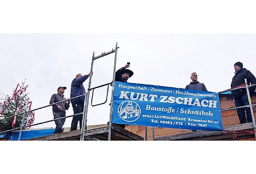 Fünf Männer stehen auf einem Gerüst an einem gemauerten Haus. An der einen Seite steht ein mit bunten Bändern geschmückter Tannenbaum. Einer der Männer hält einen Glas in die Höhe. Die anderen schauen zu ihm.