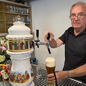 An einem Bierzapfhahn aus Porzellan mit Bemalungen und goldenen Flächen steht ein Mann und zapft ein Bier. Er schaut in die Kamera.