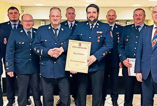 Eine Gruppe Männer, alle in ihrer Feuerwehruniform stehen in einer Gruppe und schauen zu uns. Sie haben dunkelblaue Hosen und Blauen Jacken an. Am rechten Rand steht Bürgermeister Timo Ehrhardt. In der Gruppenmitte hält ein Mann eine Urkunde in der einen Hand mit der anderen Hand gratuliert er dem Ehrenkommandanten.