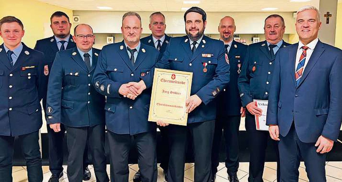 Eine Gruppe Männer, alle in ihrer Feuerwehruniform stehen in einer Gruppe und schauen zu uns. Sie haben dunkelblaue Hosen und Blauen Jacken an. Am rechten Rand steht Bürgermeister Timo Ehrhardt. In der Gruppenmitte hält ein Mann eine Urkunde in der einen Hand mit der anderen Hand gratuliert er dem Ehrenkommandanten.