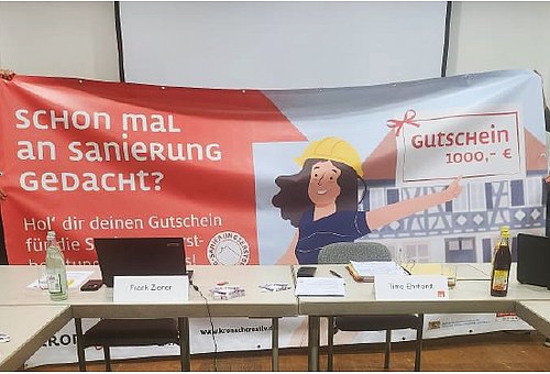 Eine Frau und ein Mann halten ein großes Banner. Darauf ist eine Zeichnung und Text zu sehen. 