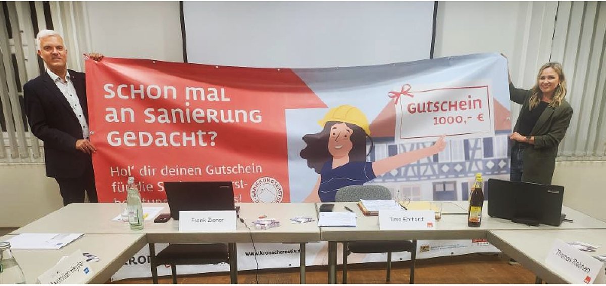 Eine Frau und ein Mann halten ein großes Banner. Darauf ist eine Zeichnung und Text zu sehen.