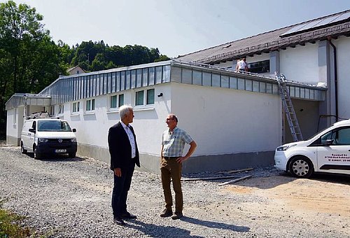 Eine Halle mit Bauumfeld. Davor stehen zwei Männer im Gespräch.