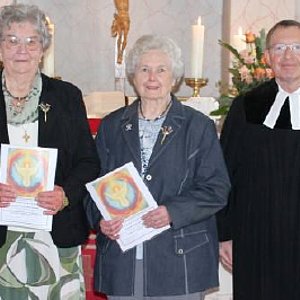 Vor einem Kirchenaltar stehen zwei Seniorinnen und ein Pfarrer in schwarzer Robe.