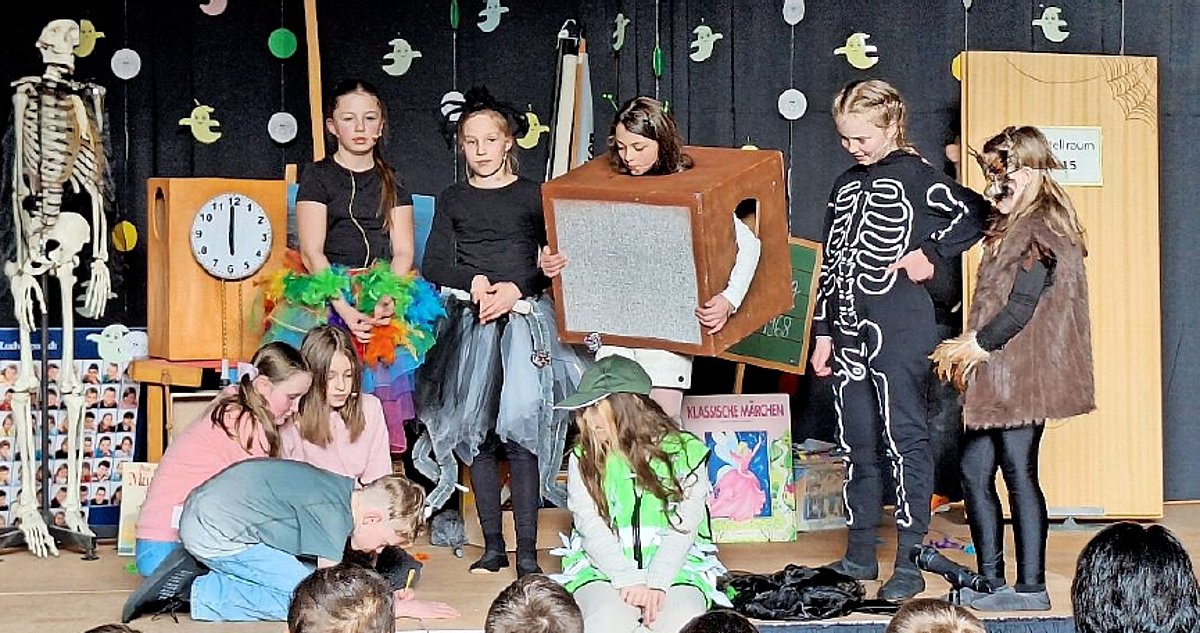 Ein Bühne in einer Halle. Darauf viele Kinder in Verkleidungen, darunter ein Skelett und Möbelstücke.
