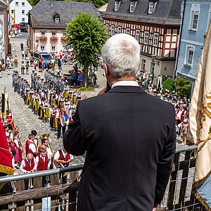 Der Bürgermeister steht auf einer Bühne und spricht zu den Menschen auf dem Marktplatz. Diese sind alle in Trachten oder Uniformen. Den Redner sieht man von hinten.