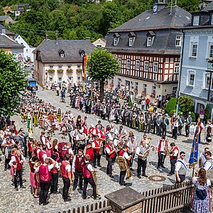 Ein Marktplatz mit Fachwerkhäusern. Auf dem Marktplatz stehen zahlreiche Gruppen von Menschen in unterschiedlichen Trachten, teilweise halten sie Fahnen oder Instrumente.