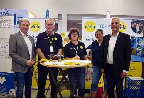 Drei Männer und zwei Frauen stehen an einem Messestand in einer Halle. Sie sind an einem Stehtisch und lächeln in die Kamera.