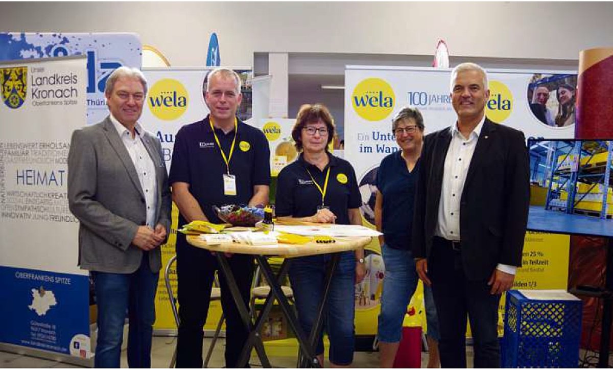 Drei Männer und zwei Frauen stehen an einem Messestand in einer Halle. Sie sind an einem Stehtisch und lächeln in die Kamera.