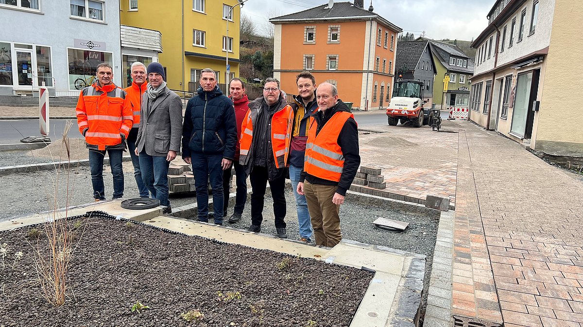Acht Männer stehen in einer Gruppe neben einer Straße. Die Flächen sind teilweise neu gepflastert. Einige der Männer tragen orange Warnwesten bzw. Warnjacken.