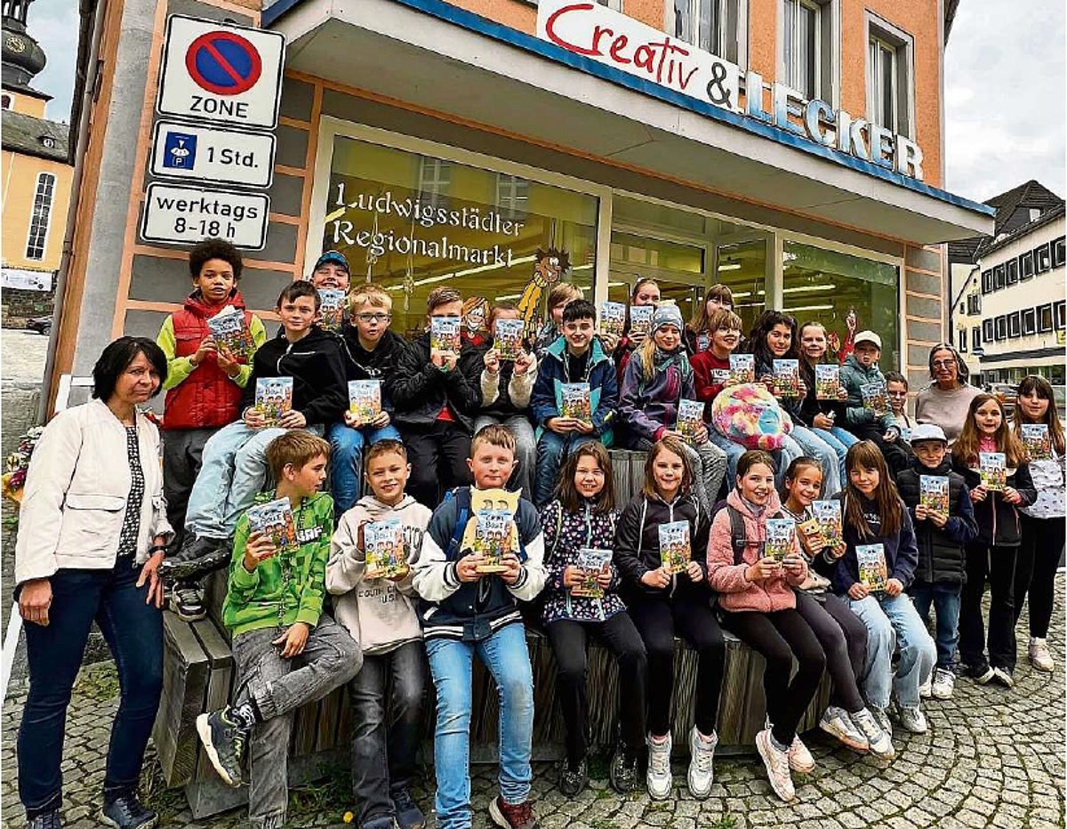 Eine große Gruppe an Schulkindern stehen in mehreren Reihen vor einem Ladengeschäft und schauen in die Kamera. Rechts und links der Gruppe stehen jeweils eine erwachsene Frau. Alle Kinder haben ein kleines Buch in der Hand.