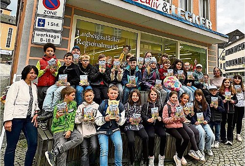 Eine große Gruppe an Schulkindern stehen in mehreren Reihen vor einem Ladengeschäft und schauen in die Kamera. Rechts und links der Gruppe stehen jeweils eine erwachsene Frau. Alle Kinder haben ein kleines Buch in der Hand. 