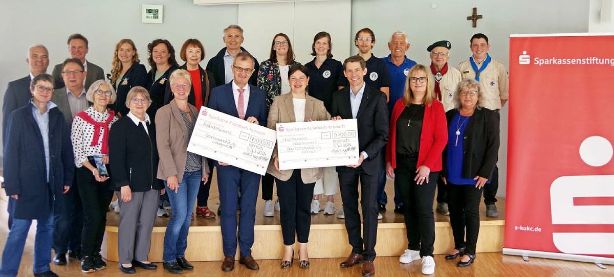 Eine große Gruppe aus Menschen bei der Spendenübergabe. In der Mitte stehen ein Mann und eine Frau, die jeweils einen großen Musterscheck in der Hand halten.