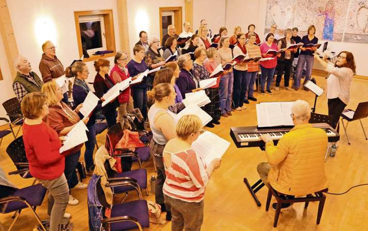 In einem großen Raum steht ein gemischter Chor. Die Gruppe ist im Profil zu sehen. Vor dem Chor steht eine Frau, die dirigiert und ein E-Piano mit einem Mann, der darauf spielt.