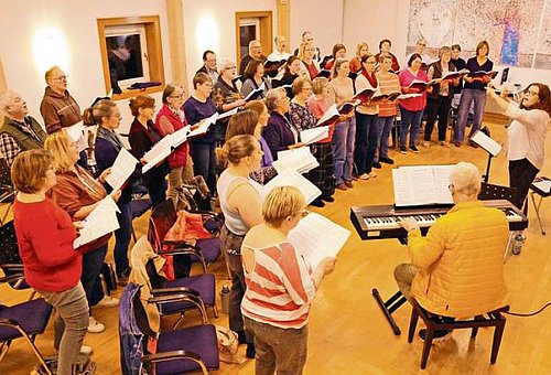 In einem großen Raum steht ein gemischter Chor. Die Gruppe ist im Profil zu sehen. Vor dem Chor steht eine Frau, die dirigiert und ein E-Piano mit einem Mann, der darauf spielt. 