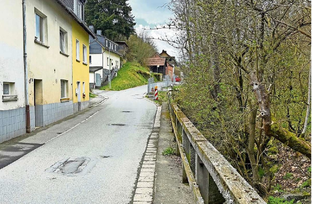 Eine enge Straße; rechts am Straßenrand eine Leitplanke und ein Bach. Auf der linken Seite eine Häuserzeile mit kleinen Häusern