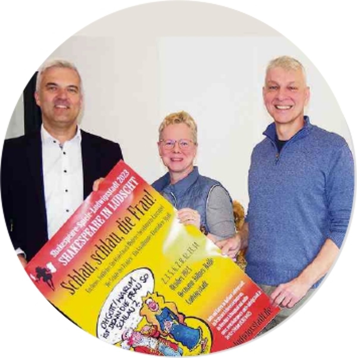 Zwischen zwei Männern steht eine Frau. Sie alle halten ein Plakat und lächeln in die Kamera.