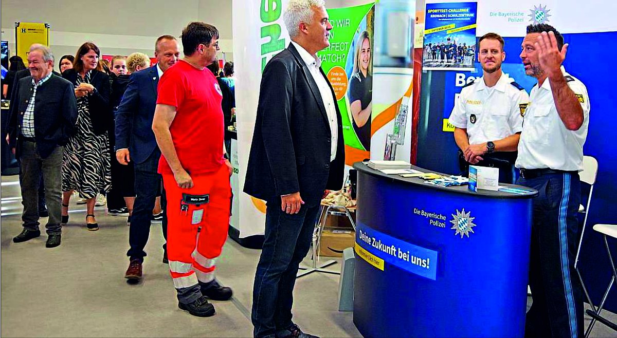 Im Vordergrund sind Bürgermeister Timo Ehrhardt mit zwei Polizeibeamten zu sehen, die sich gegenüberstehen und unterhalten. Im Hintergrund des Bildes sind mehrere Personen zu erkennen, die sich verschiedene Stände der work@ludscht anschauen.