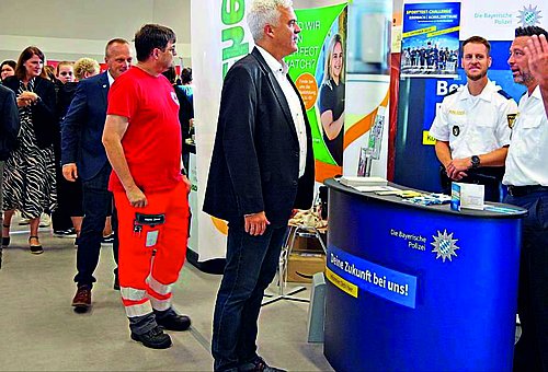 Im Vordergrund sind Bürgermeister Timo Ehrhardt mit zwei Polizeibeamten zu sehen, die sich gegenüberstehen und unterhalten. Im Hintergrund des Bildes sind mehrere Personen zu erkennen, die sich verschiedene Stände der work@ludscht anschauen.