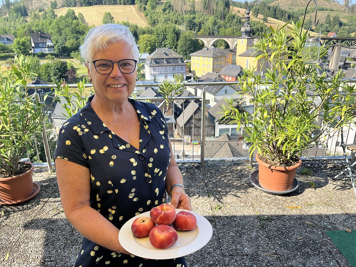 Auf einer Terrasse steht eine Frau in einem dunkelblauen Sommerkleid. In ihren Händen hält die einen Teller mit Pfirsichen und sie lächelt in die Kamera. Im Hintergrund ist eine Kleinstadt zu sehen.
