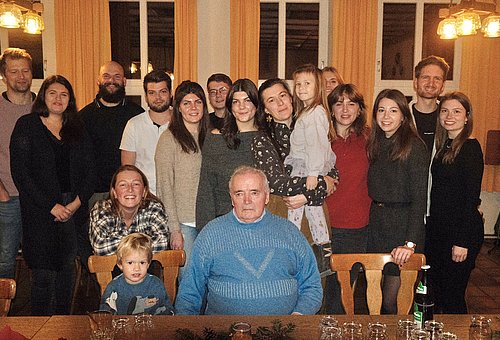 Ein alter Mann in einem blauen Pullover sitzt an einem Tisch. Dahinter stehen viele junge Leute in bunter Kleidung. Neben ihm sitzt ein Kleinkind, sein Urenkel. 