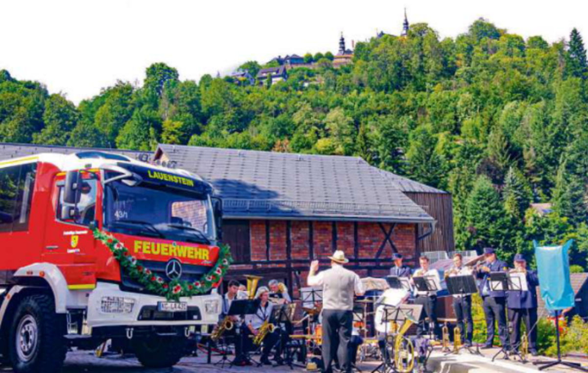 Das Foto zeigt das neue festlich geschmückte Feuerwehrfahrzeug und Musiker, die gerade auf ihren Instrumenten spielen.