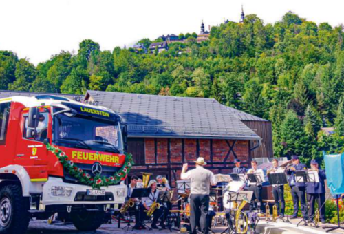 Das Foto zeigt das neue festlich geschmückte Feuerwehrfahrzeug und Musiker, die gerade auf ihren Instrumenten spielen.