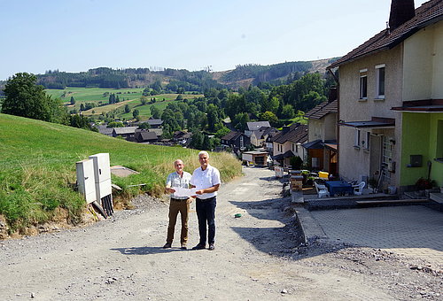 Eine unbefestigte Straße auf der einen Seite gesäumt von Wohnhäusern auf der anderen Straßenseite grüne Wiese. Auf der unbefestigten Straße stehen zwei Männer und halten ein Papier in den Händen. Sie lächeln in die Kamera.