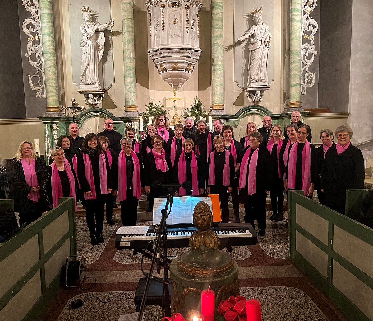 Eine Gruppe aus mehrheitlich Frauen und ein paar Männern stehen in mehreren Reihen hintereinander. Alle sind schwarz gekleidet, die Frauen zusätzlich mit einem pinkfarbenen Seidenschal. Der Chor steht im Inneren einer Kirche, alle schauen in die Kamera.