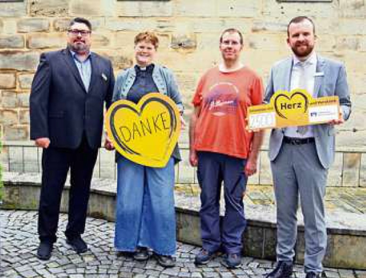 Vor einer hellen Steinmauer stehen vier Personen, darunter eine Frau. Alle tragen Alltagskleidung. Die Frau hält ein großes gelbes Herz aus Pappe in den Händen. Der Mann ganz rechts hat einen übergroßen Scheck ebenfalls mit einem gelben Herz in seiner Händen. Alle lächeln in die Kamera.